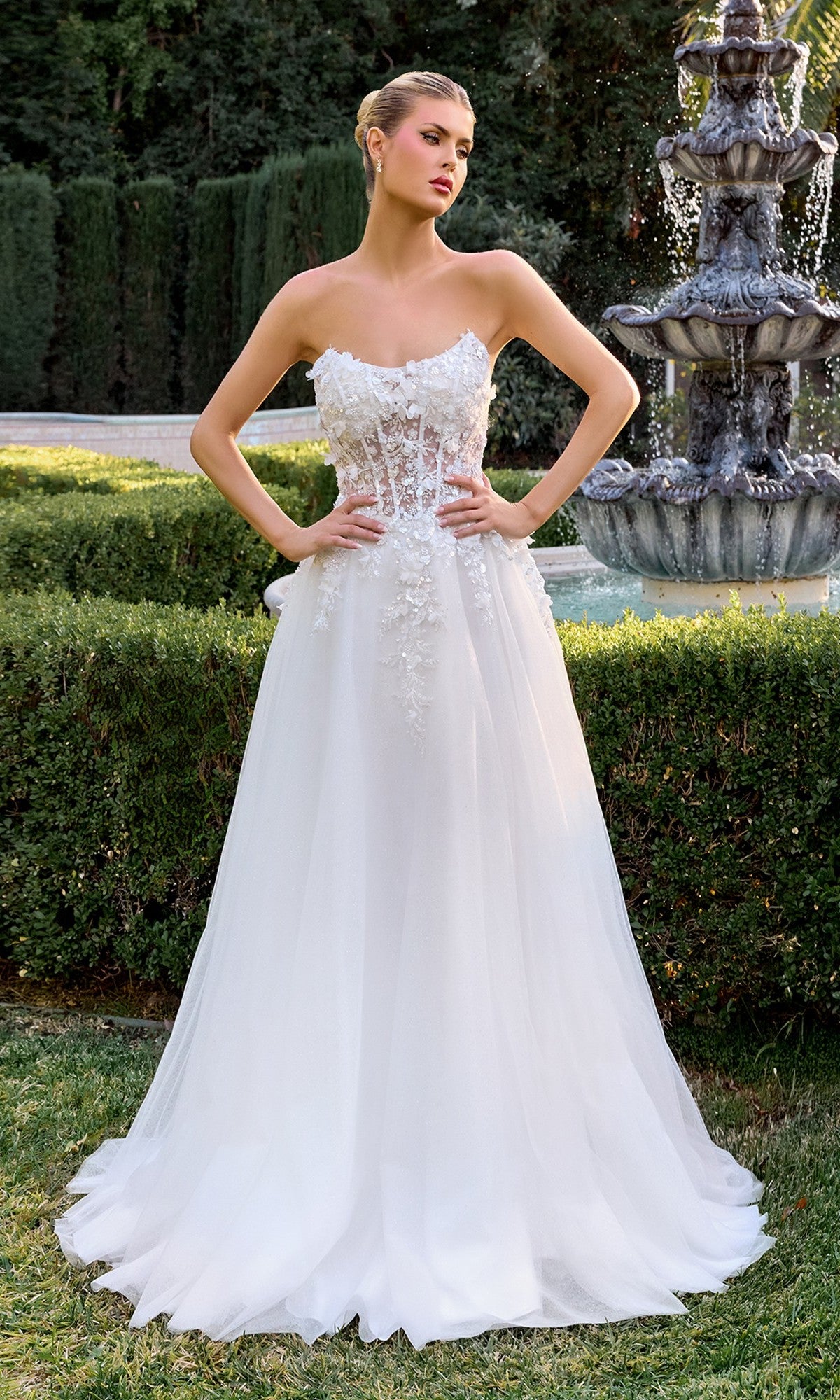 A woman poses outdoors by a fountain wearing the Ladivine C174W White Formal Dress, featuring a strapless corset bodice, floral lace details, and a tulle skirt.